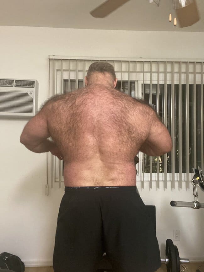 Brad Kalvo posa para fotos de costas exibindo suas costas peludas, usando uma bermuda preta.